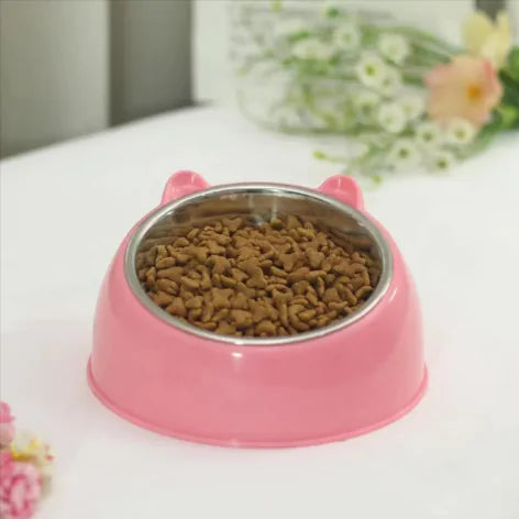 Pink pet bowl with dry food on a white surface, flowers in the background