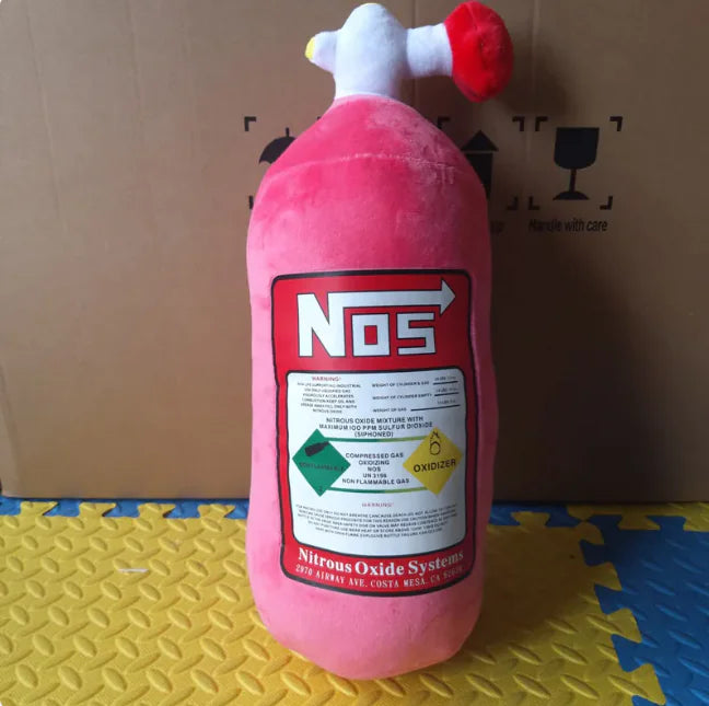 Pink plush toy resembling a NOS bottle on a textured surface with a cardboard box in the background.