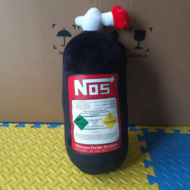 Plush toy shaped like a NOS bottle on a textured surface with a cardboard background
